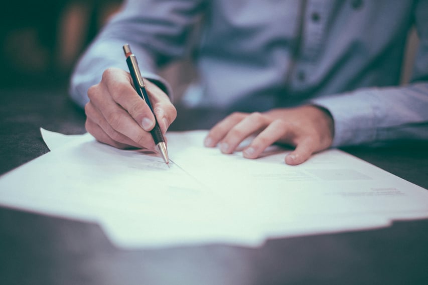 A person in a blue shirt writes with a pen on white paper at a desk, conveying focus and concentration in a professional or academic setting.