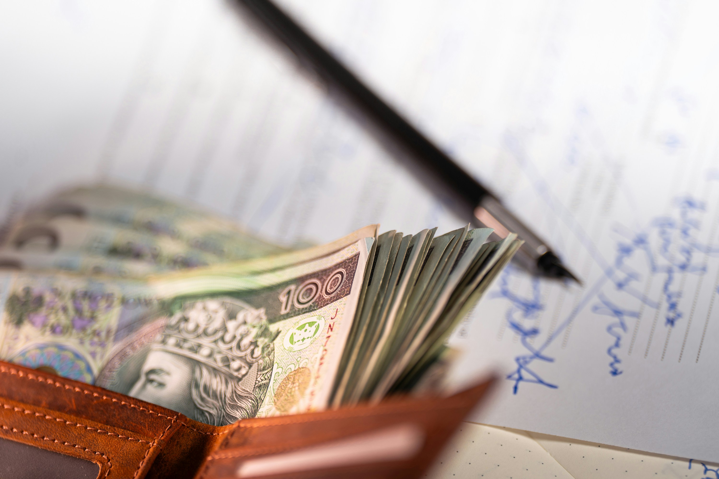 A brown wallet filled with Polish zloty banknotes rests on handwritten documents, alongside a pen. The image conveys a sense of finance and transaction.