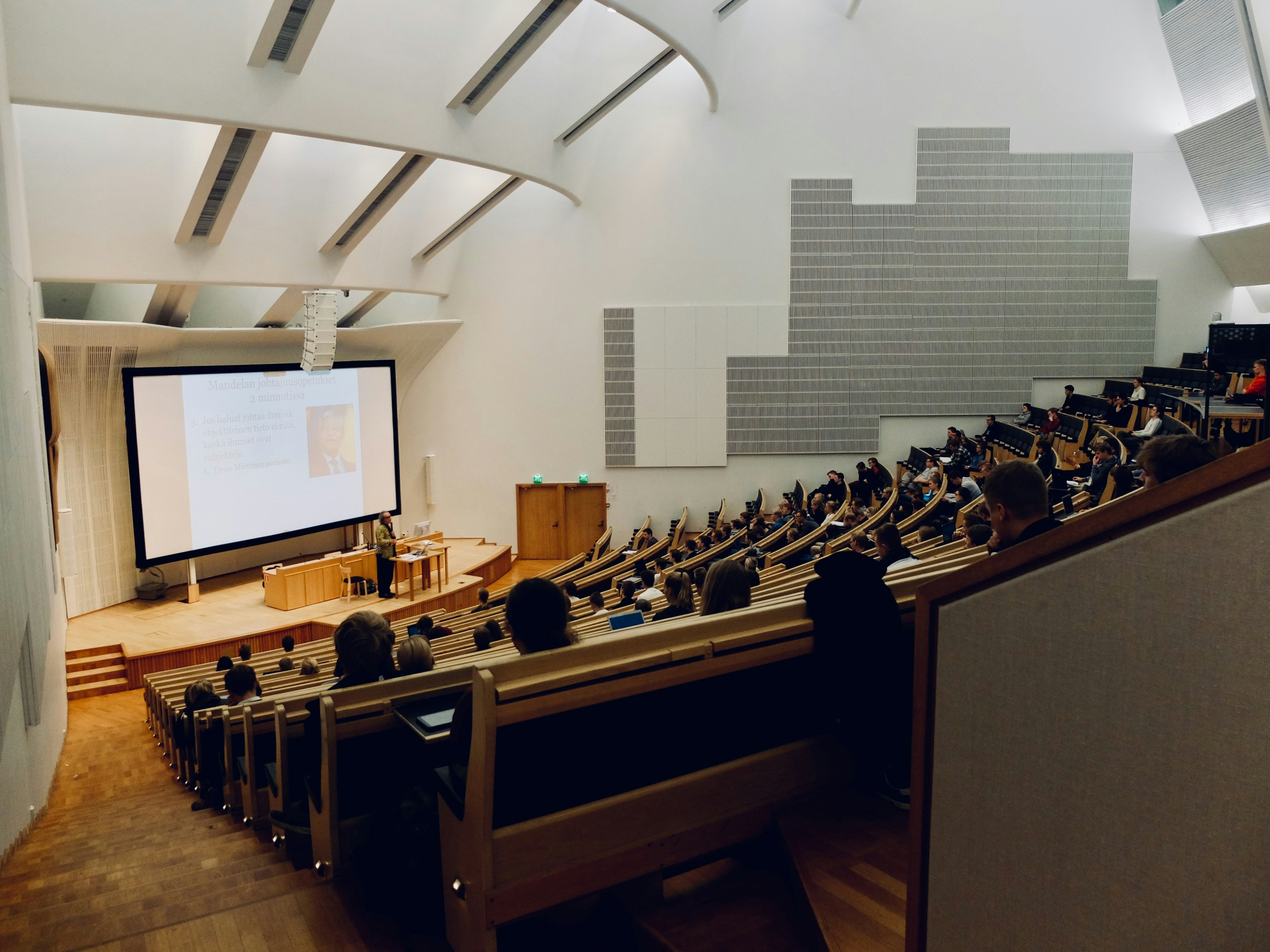A modern lecture hall with a sloped ceiling features rows of seated people facing a lecturer at a podium. A large projection screen displays content.