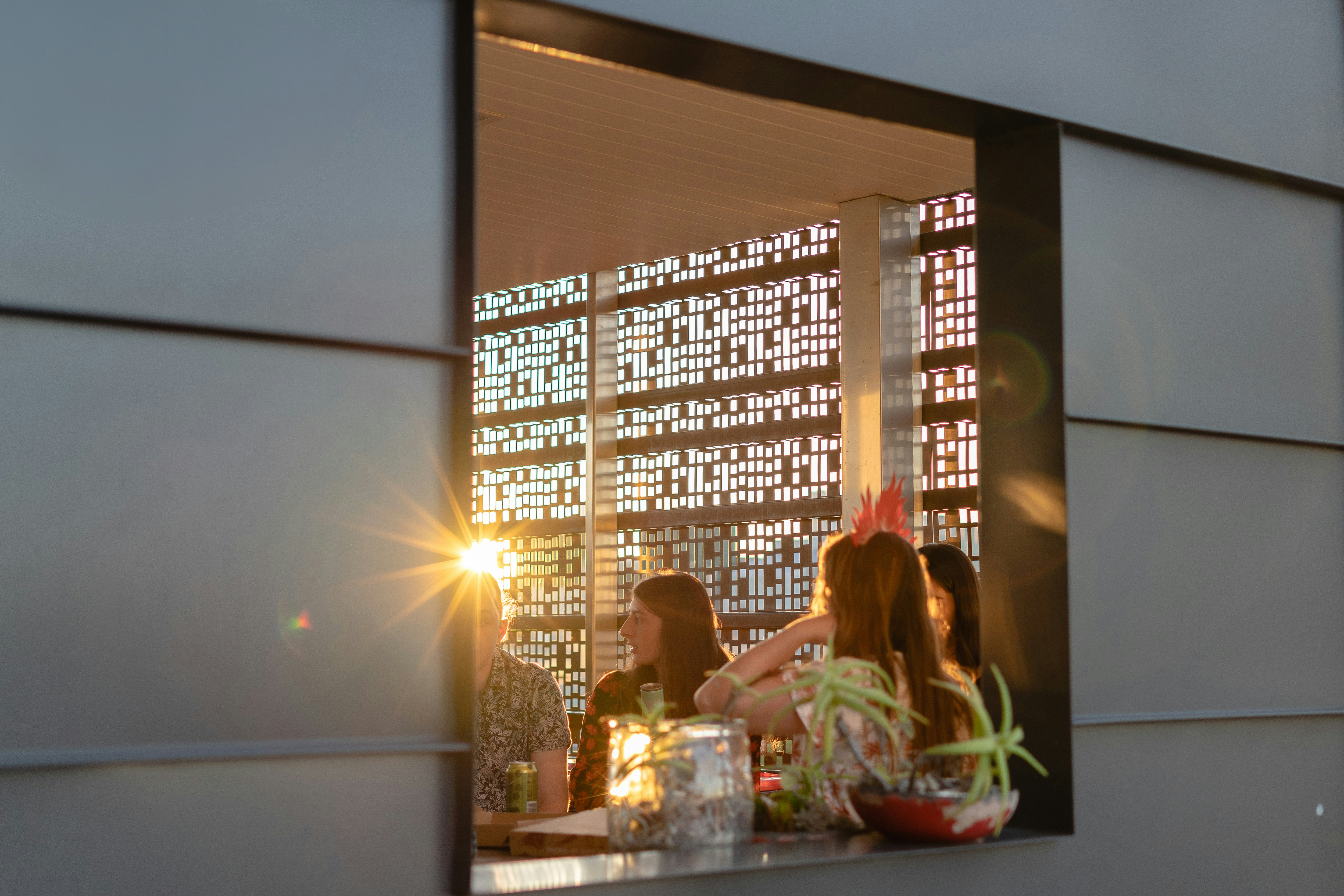 View through a window of people gathered around a table at sunset, with warm light and decorative patterned walls creating a relaxed atmosphere.