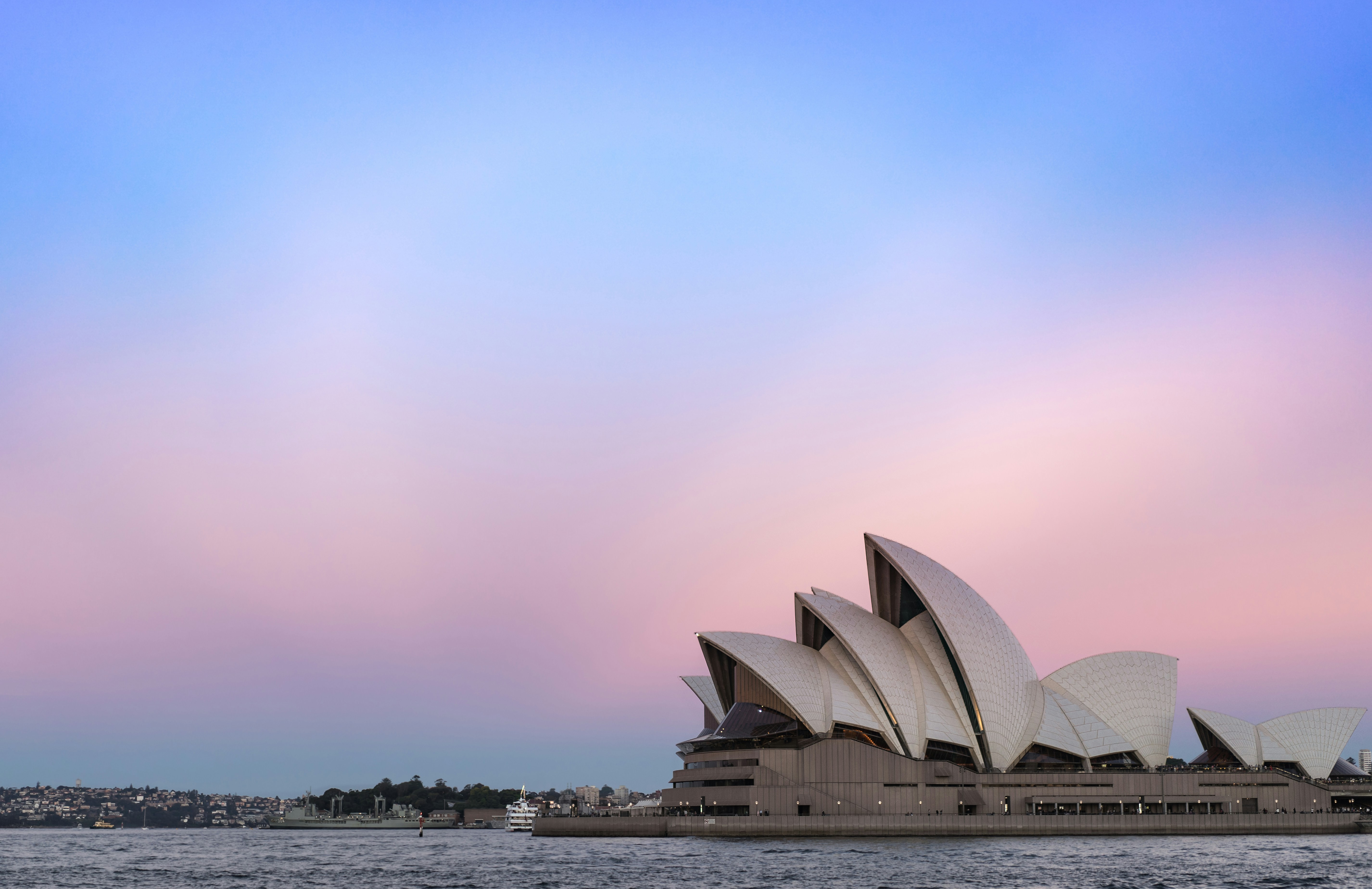 Sydney Opera House at dusk with its iconic shell-like design against a pastel pink and blue sky. Calm water foreground creates a serene atmosphere.