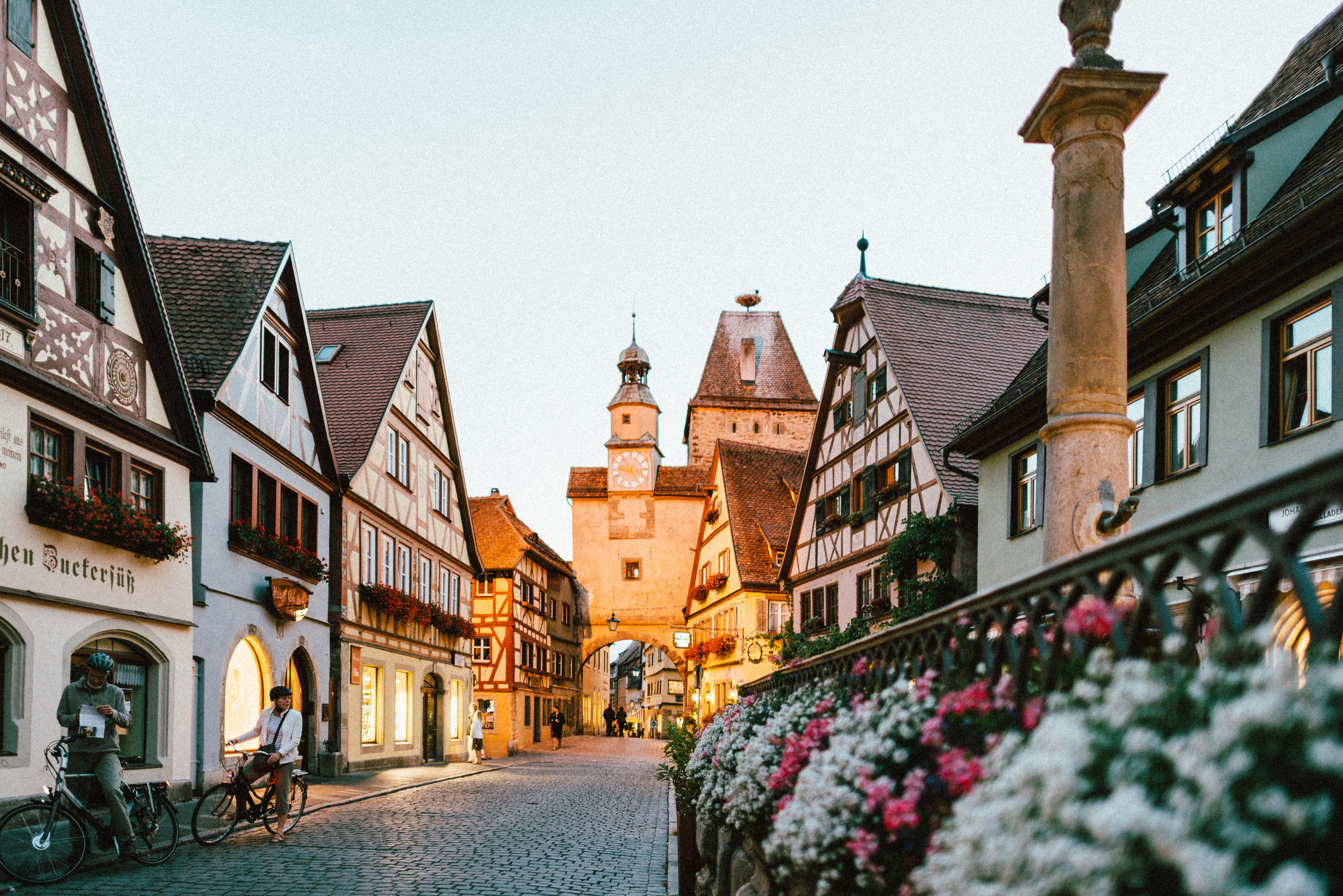 Quaint cobblestone street flanked by half-timbered houses, clock tower, bicycles, and vibrant flowers at sunset.