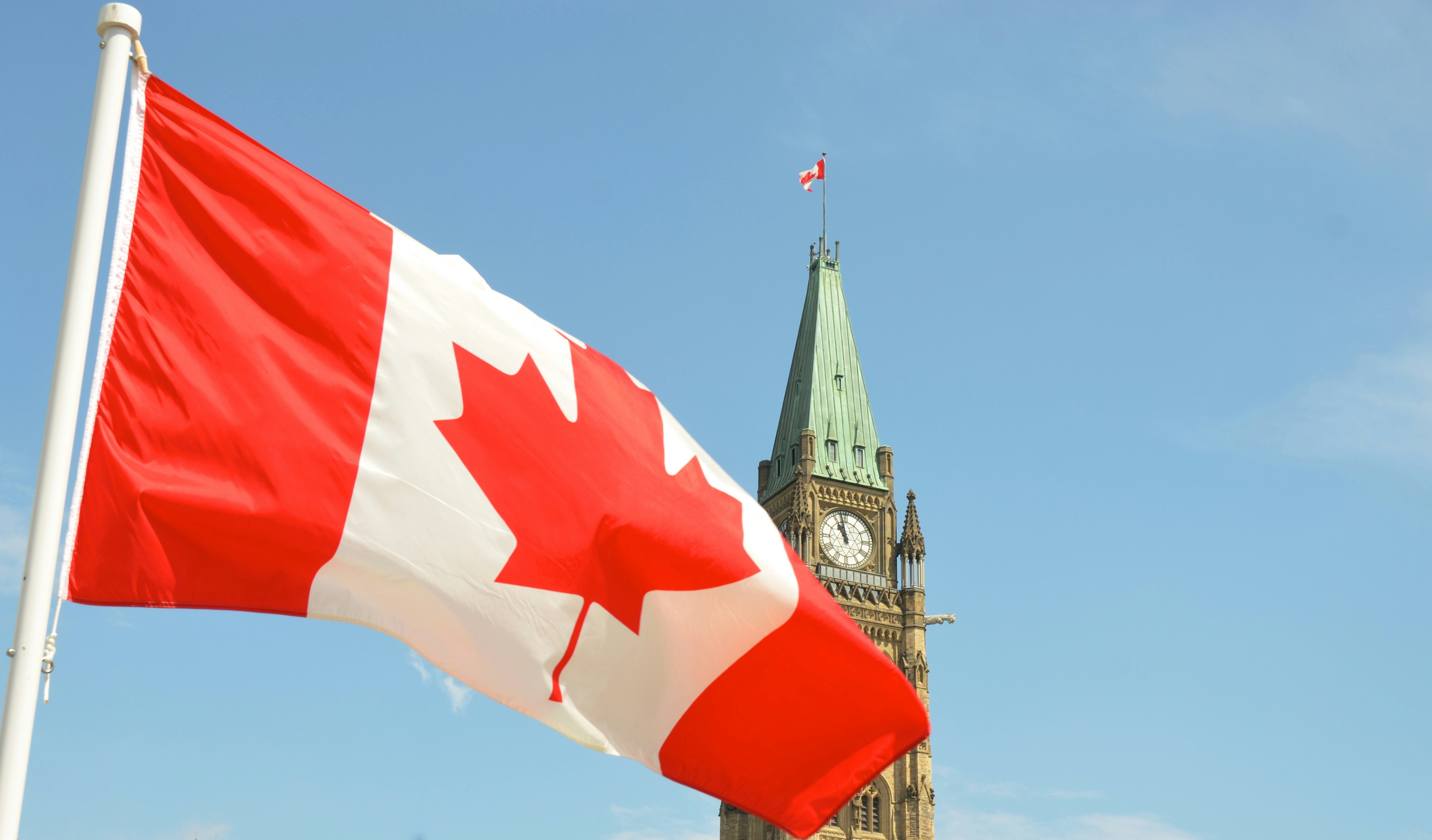 Canadian flag waving prominently against a clear blue sky, with the Peace Tower of Parliament in Ottawa visible in the background, conveying national pride.